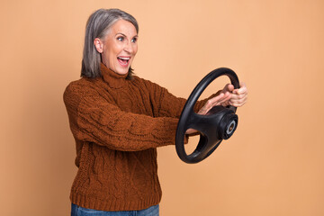 Joyful elderly woman gestures energetically holding steering wheel, beige background