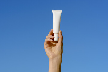 Female hand holding a white thin mockup tube of cream or lotion or serum on a blue sky background....