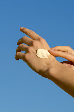 Vertical image of female hands applying a sample of swatch cream on the skin to moisturize and smooth the skin on a blue sky background. Concept of smear, care cosmetics, beauty products.