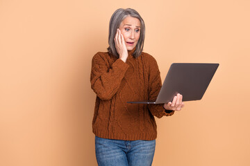 Stylish mature woman looking surprised with laptop against beige background