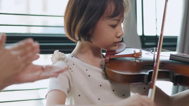 Asian young girl practices violin with focused dedication, guided by a supportive teacher showing music education, classical music, and the bond between student and mentor for nurturing talent.