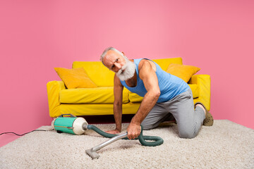 Senior man vacuuming carpet with yellow sofa and pink background in a vibrant interior setting