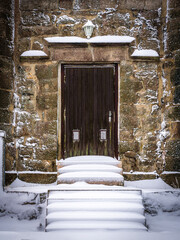 A rustic wooden door in a stone surround surrounded by snow-covered steps and decorative flower pots
