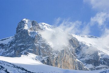 A snowy rocky mountain peak with wind blowing snow and a blue sky near a ski resort in the French or Swiss Alps in the winter
