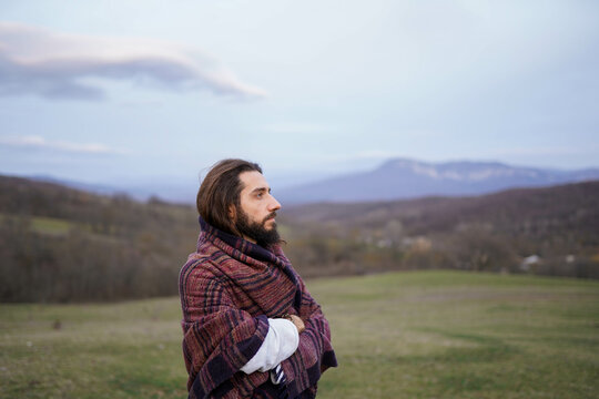 Side portrait of bearded man wrapped in ethnic blanket looking into distance on green hills