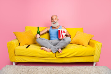 Obraz premium Elderly man relaxing on a vibrant yellow sofa with popcorn and drink against pink background