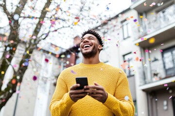 A young man wearing a yellow sweater enjoys a moment of laughter while checking his phone as confetti falls around him