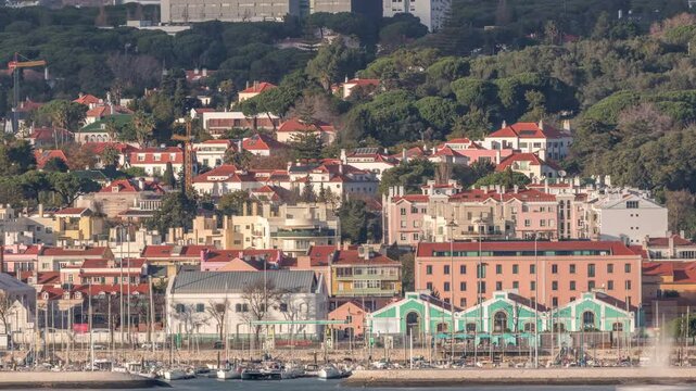 Aerial timelapse of Marina Doca do Bom Sucesso in Lisbon, Portugal, once a naval harbor and now a leisure port. Colorful houses with red roofs line the waterfront against the Tagus River backdrop.