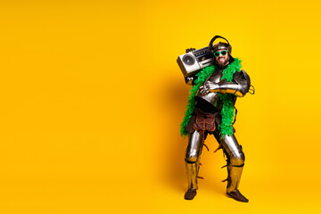 Young man in knight costume with boombox and feather boa on yellow background