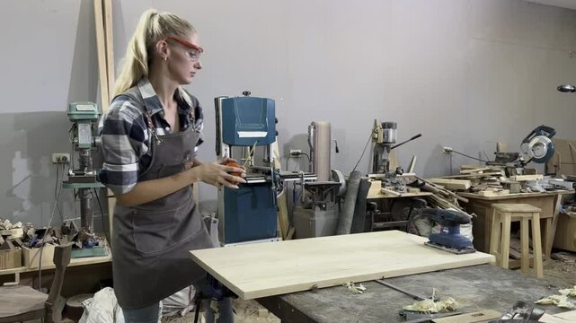 Craftswoman at Work: A focused young woman wearing safety glasses and an apron meticulously works on a wooden surface with a sander in a woodworking shop, surrounded by various tools and equipment.