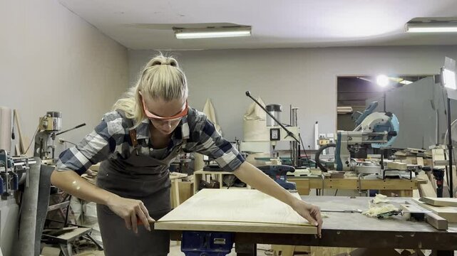 Carpentry Craftsmanship: A focused woman with blonde hair tied back, wearing safety glasses and overalls, meticulously works on a piece of wood in a workshop, surrounded by tools and machinery.