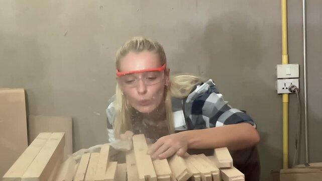 A Carpenter Blowing Dust Off Wood: A focused carpenter, wearing safety goggles, blows sawdust off a pile of wooden planks in a workshop, highlighting the hands-on nature of craftsmanship.