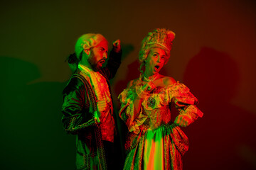 Classically styled couple in vibrant costumes posing dramatically against a red and green lit dark...