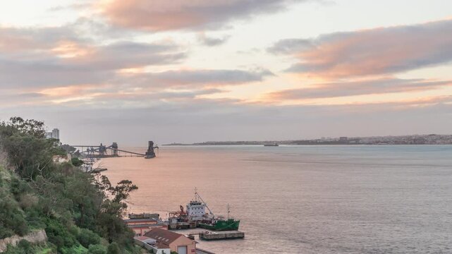 Aerial timelapse from a hill near Porto Brandao Ferry Station on the Tagus River in Lisbon. Dramatic sunset with colorful clouds, a ship floating on water and Terminal Portuario Trafaria in background