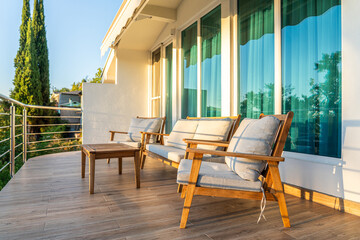 Modern outdoor balcony with wooden chairs, cushions, coffee table, and glass doors in a sunny setting