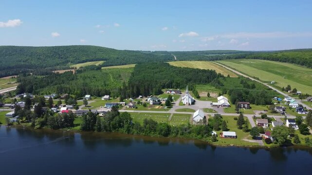 Drone footage overlooking the peaceful village of Saint‑Z&eacute;non‑du‑Lac‑Humqui, with its church, lakeside cemetery, wooded hills, and agricultural fields in the Matapedia Valley, Quebec, Canada.