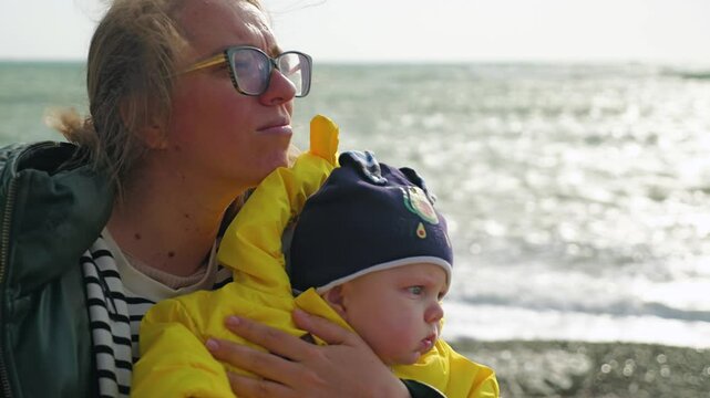 Close-up of mother and child with ruddy cheeks with bright blue eyes and yellow jacket on the beach. The wind is ruffling the hair of a woman in glasses who is holding her son in her arms
