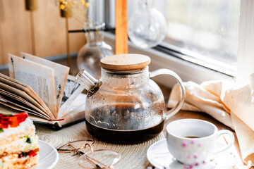 a piece of cake with buttercream on pieces of multi-colored jelly on a white saucer on a wooden tray under the sun's rays, espresso coffee in a white cup, a transparent teapot with coffee, flowers, a 