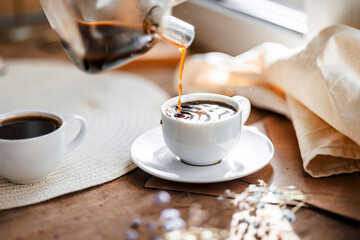 espresso coffee is poured into a white cup on a white saucer on a wooden tray under the sun's rays, a transparent teapot with coffee, flowers, a book, morning coffee, a drink, a light composition