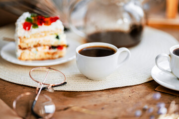 a piece of cake with buttercream on pieces of multi-colored jelly on a white saucer on a wooden tray under the sun's rays, espresso coffee in a white cup, a transparent teapot with coffee, flowers