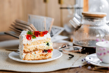 a piece of cake with buttercream on pieces of multi-colored jelly on a white saucer on a wooden tray under the sun's rays, espresso coffee in a white cup, a transparent teapot with coffee, flowers