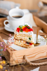 a piece of cake with buttercream on pieces of multi-colored jelly on a white saucer on a wooden tray under the sun's rays, espresso coffee in a white cup, a transparent teapot with coffee, flowers