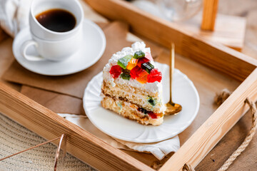 a piece of cake with buttercream on pieces of multi-colored jelly on a white saucer on a wooden tray under the sun's rays, espresso coffee in a white cup, a transparent teapot with coffee, flowers