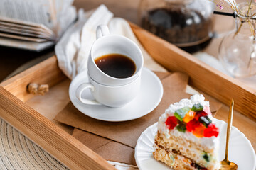 a piece of cake with buttercream on pieces of multi-colored jelly on a white saucer on a wooden tray under the sun's rays, espresso coffee in a white cup, a transparent teapot with coffee, flowers