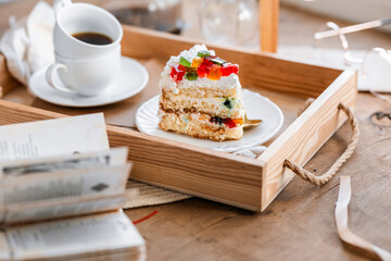 a piece of cake with buttercream on pieces of multi-colored jelly on a white saucer on a wooden tray under the sun's rays, espresso coffee in a white cup, a transparent teapot with coffee, flowers