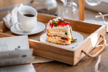 a piece of cake with buttercream on pieces of multi-colored jelly on a white saucer on a wooden tray under the sun's rays, espresso coffee in a white cup, a transparent teapot with coffee, flowers