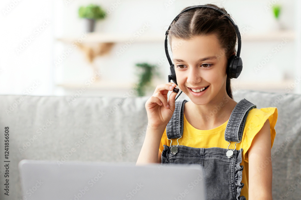 Poster Kids And Technology Concept. Portrait of smiling girl sitting on couch at table and using laptop computer, wearing and touching headset talking in microphone having video call or playing online game