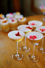 Several cocktail glasses filled with a light-colored drink topped with a frothy layer. Each drink is garnished with a colorful flower, with some flowers being red and others pink