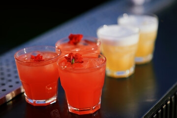 Five glasses of drinks placed on a dark surface. Three glasses contain a red-colored drink with ice cubes, each garnished with a small red flower