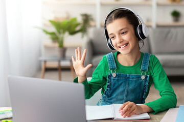Distance Learning. Cute teenage girl in headset having video lesson using laptop in living room, studying remotely, enjoying online education, waving hand at pc web camera