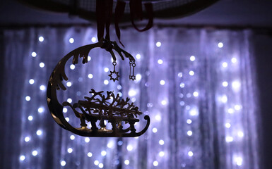 Mysterious holiday photo: on the background of a window with a shining garland hangs a golden pendant - a Christmas tree on a sleigh with golden stars hanging from it. The background is blurred.