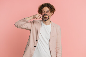 Happy Caucasian man holding magnifier glass on teeth, looking at camera, showing funny smiling silly face mouth. Health care, hygiene, stomatology. Curly haired young guy isolated on pink background