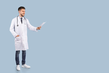 Smiling Male Doctor Recommending Medical Treatment Posing Holding Paper Sheet Standing Over Blue Studio Background, Wearing White Uniform. Panorama With Free Space, Full Length