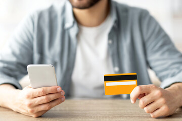 Shopping at home. Closeup of young guy holding credit card and smartphone, making online purchase at table indoors. Young Caucasian man buying goods on web, cropped view