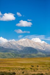 Majestic mountain range under a clear blue sky with fluffy clouds and lush green hills at midday