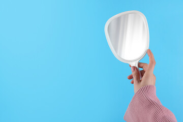 Woman holding handheld mirror on light blue background, closeup. Space for text