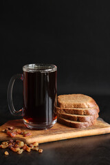Mug of tasty kvass, bread and raisins on a dark background. Glass mug of kvass on a wooden board, close-up. Dark kvass with ingredients on a black background. Refreshing drink with slices of bread