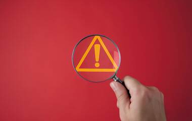 Cybersecurity Alert: Magnifying Glass Reveals Glowing Warning Sign on Red Background. Hand...