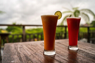 On a wooden table in Bali, two vibrant drinks, an orange one garnished with a lime slice and a red...