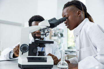 Dark-skinned schoolgirl in a lab coat looking into microscope and looking involved