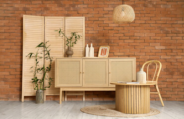 Wooden chest of drawers, table, chair and folding screen near brick wall in stylish interior of...