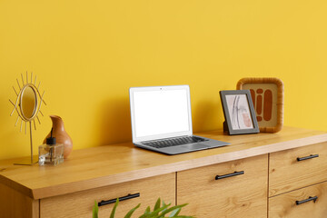 Blank laptop with frames on wooden commode near yellow wall