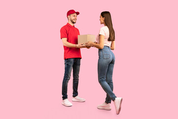 Delivery Courier Guy Giving Parcel Box To Woman Standing Over Yellow Studio Background. Post Packages Delivering, Parcels Transportation Service Concept. Full Length