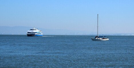 A ferry and sailboat share the calm waters of the San Francisco Bay on a sunny day. The Golden Gate Bridge can be seen in the distance.b