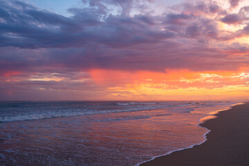 Fototapeta premium Waves roll in under a vibrant, dramatic sky painted with deep sunset hues.