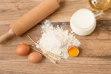 Heap of flour with eggs, wheat spikelets, jug of milk and rolling pin for preparing dough on wooden background